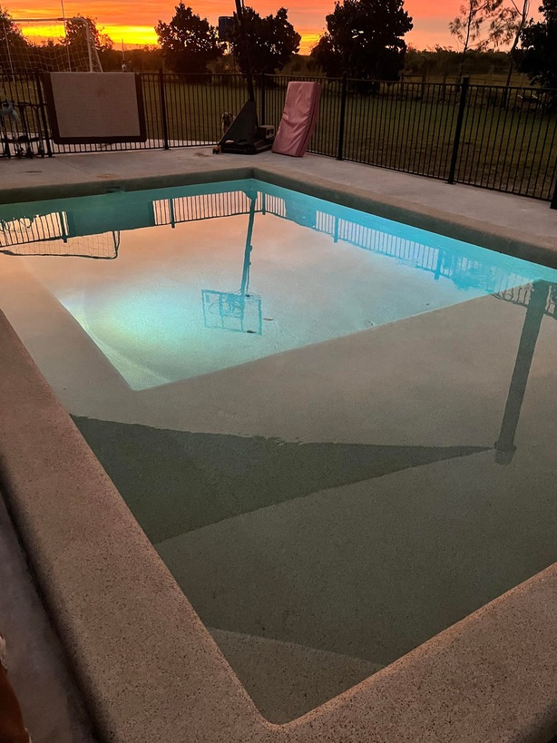 Pool lit up at dusk with a sunset sky and pool fence behind