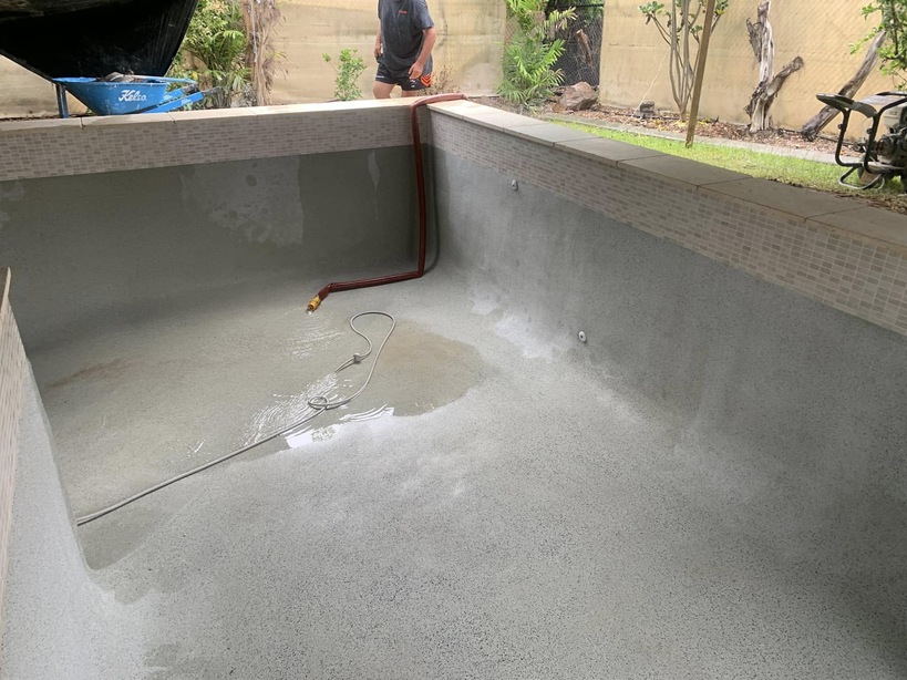 Empty pool shell with a grey pebble interior, hose on the floor and a worker on the edge