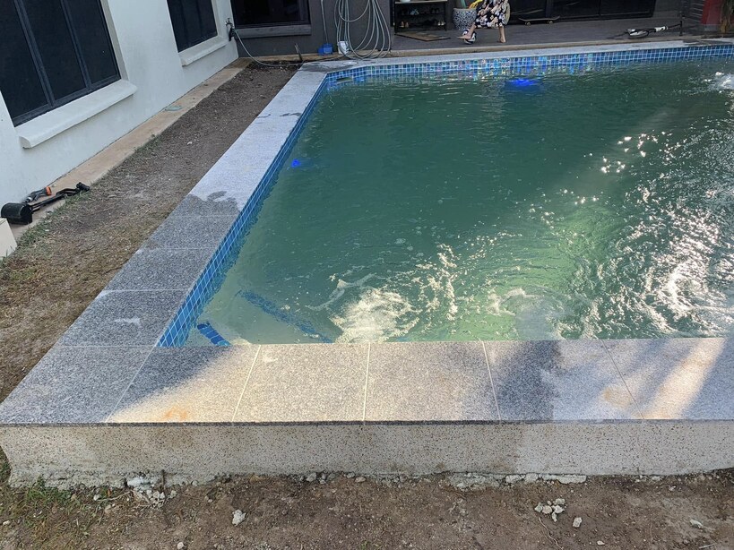 Raised pool with granite coping and jets running beside a white house